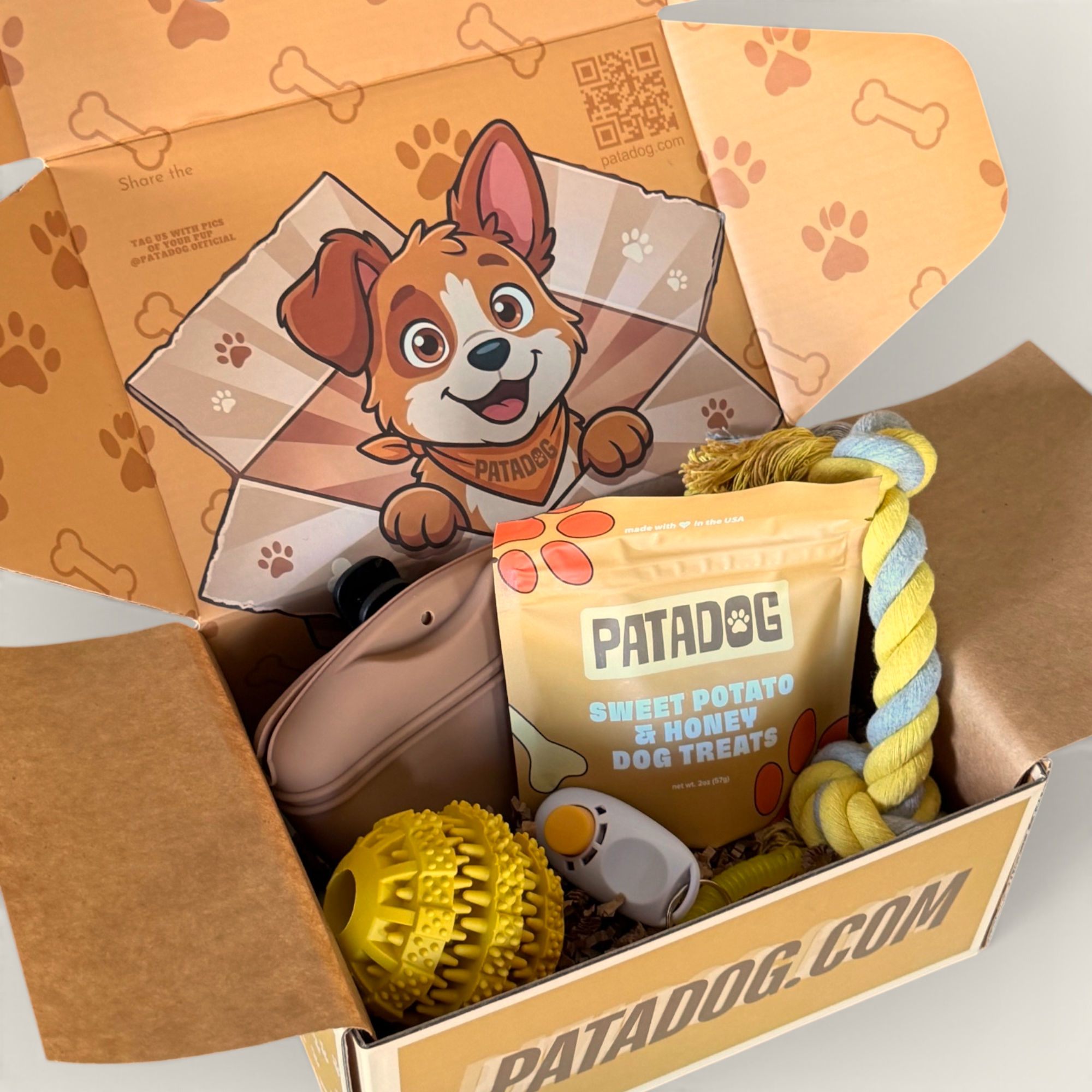 An open Patadog box filled with dog treats and toys, including a bag of Sweet Potato & Honey Dog Treats, a yellow squeaky ball, a rope toy, clicker, and a brown plush toy, set against a clean white background.