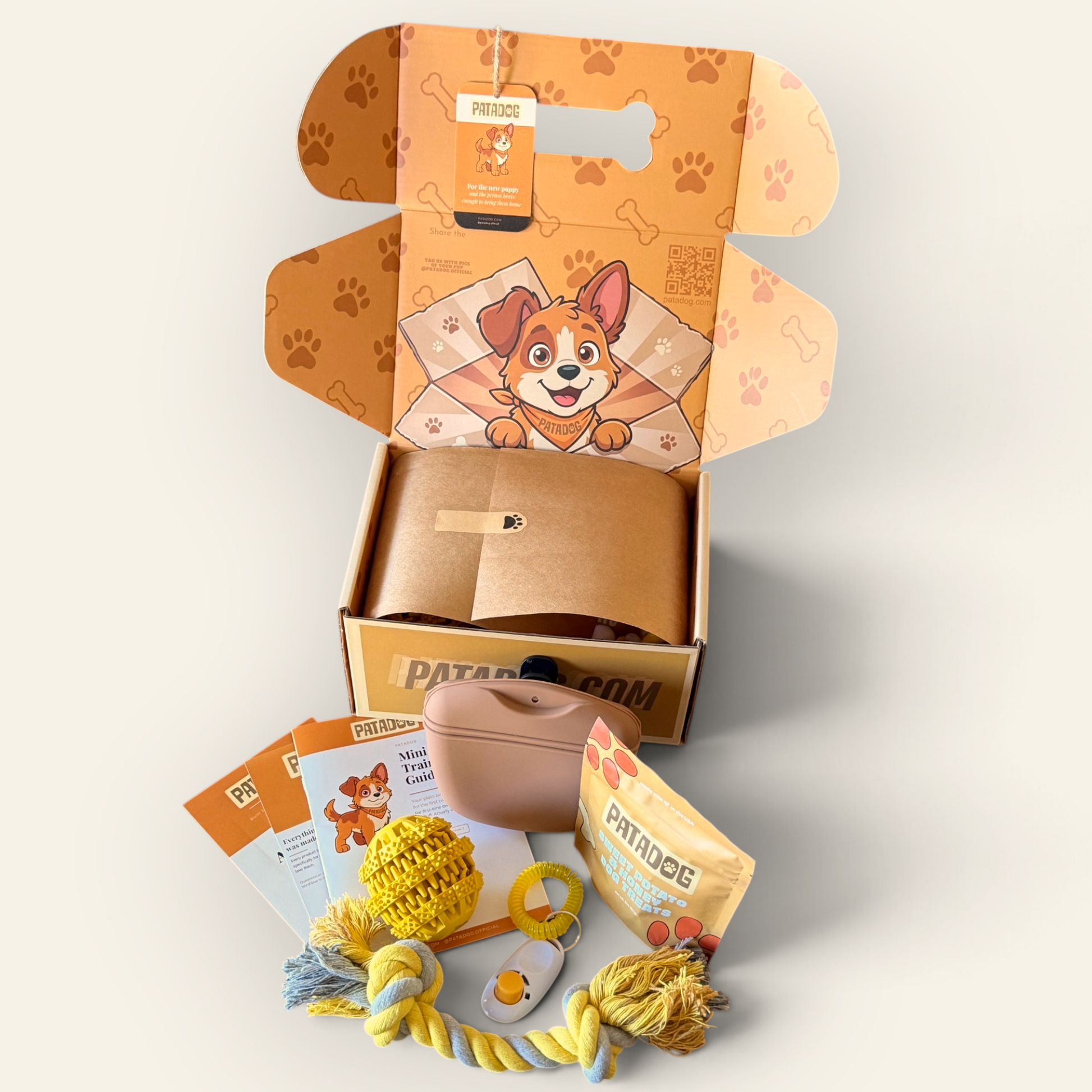 An open Patadog pupoy box displayed on a light beige background, showing a brown tissue insert with a cartoon dog illustration, a small tan pet case, a clicker, training guide and several pet toys and treats arranged in front. The packaging features orange paw-print patterns and the Patadog brand name.